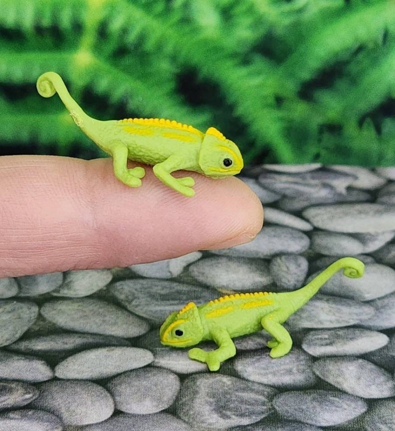 May include: Two small green and yellow toy chameleon figurines. The chameleon on the left is on a finger, while the chameleon on the right is on a bed of small grey rocks.