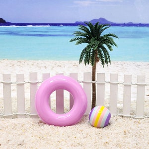 May include: A pink inflatable ring and a colorful striped beach ball on a white picket fence with a palm tree in the background.