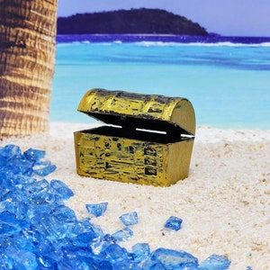 May include: A small, gold-colored treasure chest with a hinged lid, open to reveal the empty interior. The chest is sitting on a white sandy beach with blue glass pieces scattered around it.