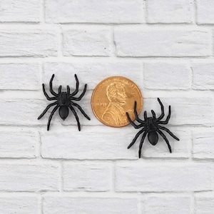May include: Two black plastic spiders, each about the size of a US penny, on a white brick background.