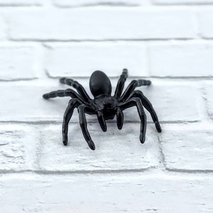 May include: A black plastic spider toy with eight legs. The spider is on a white brick background.