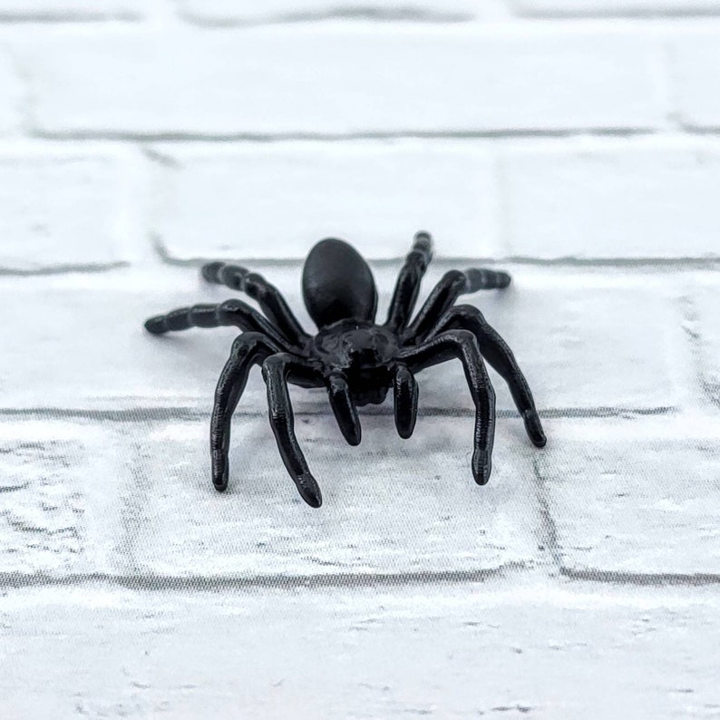 May include: A black plastic spider toy with eight legs. The spider is on a white brick background.