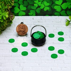 May include: A miniature black cauldron filled with green shamrock coins, with additional shamrock coins scattered on a white brick surface. A copper penny is displayed next to the cauldron. Green shamrock decorations are in the background.