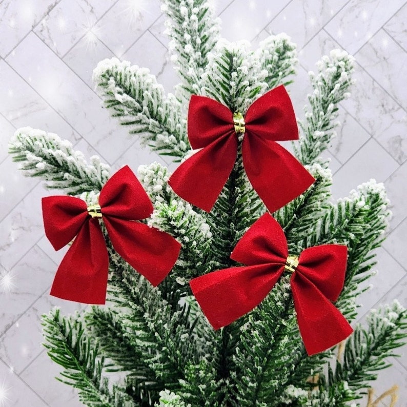 May include: Three red velvet bows with gold centres are tied to a green Christmas tree.