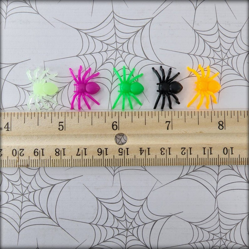 May include: Five plastic toy spiders in different colors: green, pink, black, orange, and glow-in-the-dark green. The spiders are arranged in a row on a white surface with a wooden ruler behind them.