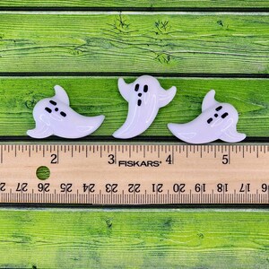 May include: Three white glass ghost ornaments with black eyes and mouths. The ghosts are approximately 1 inch tall and are sitting on a wooden ruler.