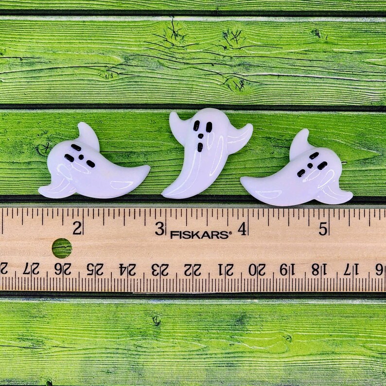 May include: Three white glass ghost ornaments with black eyes and mouths. The ghosts are approximately 1 inch tall and are sitting on a wooden ruler.