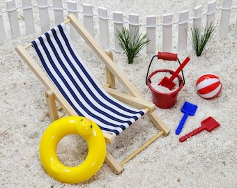 May include: A blue and white striped beach chair with a yellow inflatable ring, a red bucket with a red shovel, and a red and white beach ball on a white sand background.