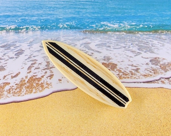 May include: A wooden surfboard with black stripes, lying on a sandy beach with blue ocean waves in the background.
