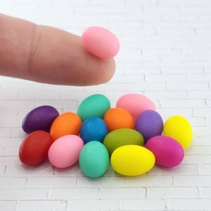 May include: A collection of miniature Easter eggs in various colors, including pink, purple, red, orange, yellow, green, and blue. The eggs are arranged in a pile on a white brick background.