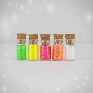 May include: Five small glass jars with cork lids filled with different colored glitter. The jars are lined up in a row from left to right: green, yellow, pink, orange, and white.