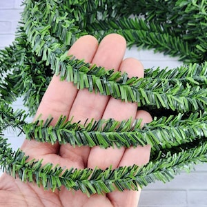 May include: Green artificial pine garland with a textured, feathery appearance. The garland is shown in a hand, demonstrating its flexibility.