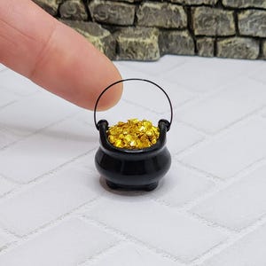 May include: A miniature black cauldron filled with gold coins, with a black wire handle. The cauldron is sitting on a white tiled surface.