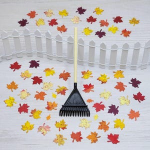 May include: A miniature black rake with a light beige handle is surrounded by colorful paper maple leaves in shades of red, orange, and yellow. A small white picket fence is in the background, creating a fall-themed scene.