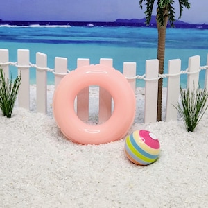 May include: A pink inflatable pool float shaped like a ring and a colorful striped beach ball on a white sandy beach.