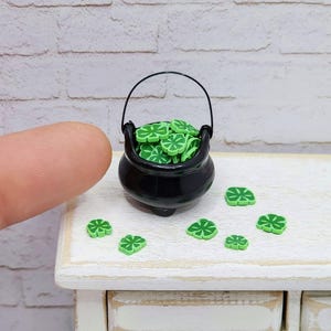 May include: A miniature black cauldron filled with green four-leaf clover charms.  There are also several charms scattered on a white wooden surface.