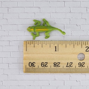 May include: A small green and yellow chameleon figurine measuring approximately 2.5 cm in length. The figurine is posed in a walking position with its legs outstretched and its tail curled.