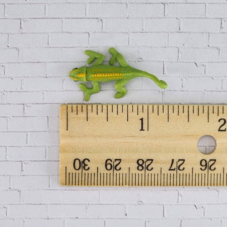 May include: A small green and yellow chameleon figurine measuring approximately 2.5 cm in length. The figurine is posed in a walking position with its legs outstretched and its tail curled.