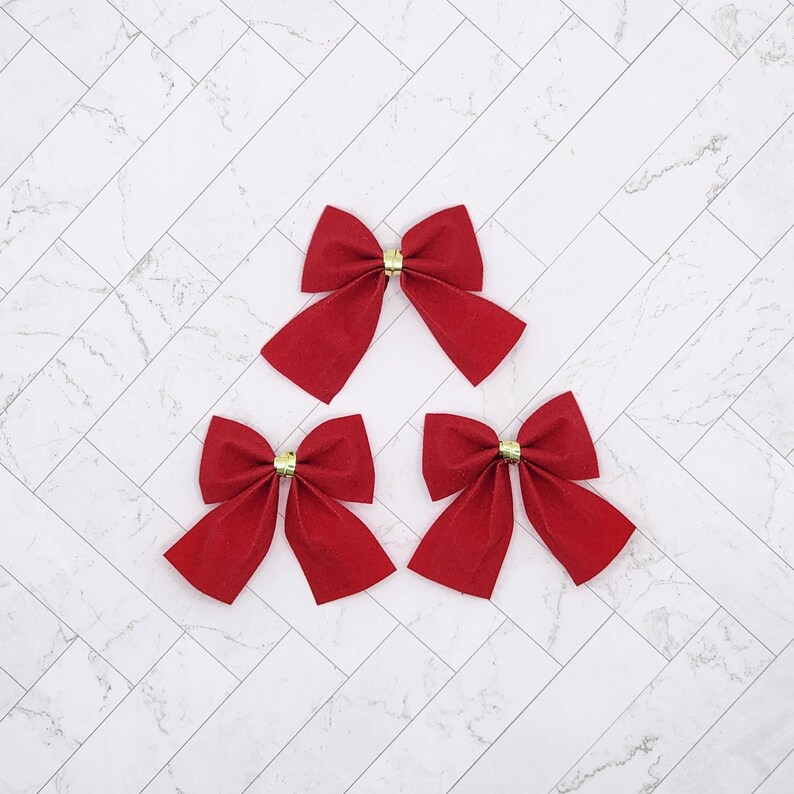 May include: Three red velvet bows with gold centres. The bows are arranged in a triangular pattern on a white marble background.