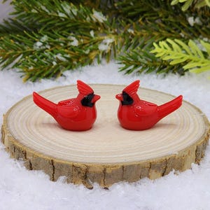 May include: Two red glass cardinal figurines with black eyes and beaks, perched on a wooden log slice. The figurines are facing each other, creating a romantic and festive scene.