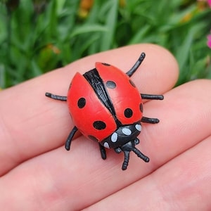 May include: A small, red ladybug figurine with black spots and a black head and legs. The ladybug is resting on a person's hand, with a blurred green background.