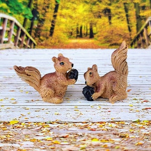 May include: Two brown squirrel figurines with black eyes and noses, holding black acorns in their paws. The squirrels are sitting on a white wooden surface.