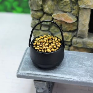 May include: A miniature black cauldron filled with gold coins. The cauldron has a handle and is sitting on a gray wooden surface.