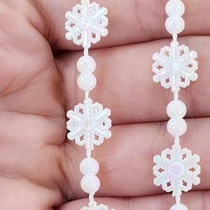 May include: White snowflake garland with iridescent glitter and white pearl beads. The garland is made of plastic and is perfect for decorating for the winter holidays.