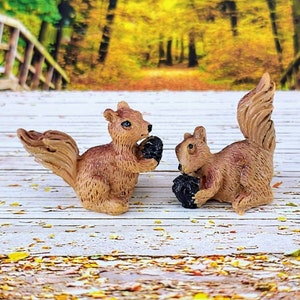 May include: Two miniature brown squirrels, each holding a dark object, possibly a nut or seed. The squirrels are positioned on a white wooden surface, with a blurred autumn-themed background of trees and foliage.