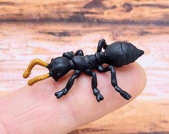 May include: A black plastic ant figurine with tan antennae is resting on a finger. The ant has six legs and a segmented body. The background is a blurred wooden surface.