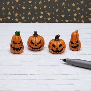 May include: Four miniature orange jack-o'-lanterns with carved faces. Each pumpkin has a unique expression and stem. The pumpkins are on a white brick surface with a starry background. A pen is in the foreground.
