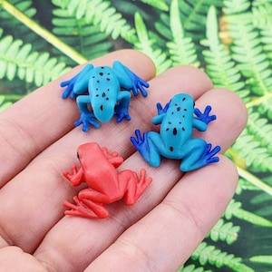 May include: Three small plastic frog figurines. One frog is blue with black spots, one is red, and one is blue with black spots. The frogs are sitting on a hand.