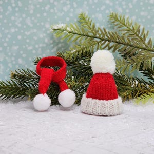 May include: A red knitted Santa hat with a white pom-pom and a white trim, and a red knitted scarf with white pom-poms, on a snowy background.