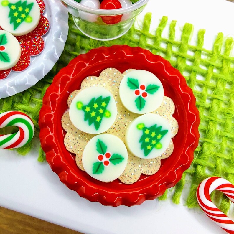 May include: A red and white plate with four white candy ornaments decorated with green Christmas trees and holly. The plate is on a green woven placemat.