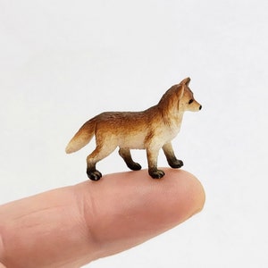 May include: A small, detailed figurine of a red fox standing on a finger. The fox has a bushy tail and a pointed snout.