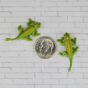 May include: Two green plastic toy lizards with yellow stripes on a white brick background. One lizard is facing left and the other is facing right. A US dime is in the centre of the image for scale.
