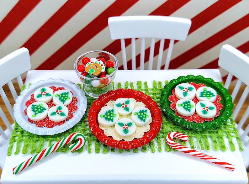 May include: A white table set with a green and brown placemat, two candy canes, a bowl of candy, and three plates of Christmas tree cookies. The cookies are white with green trees and red holly.