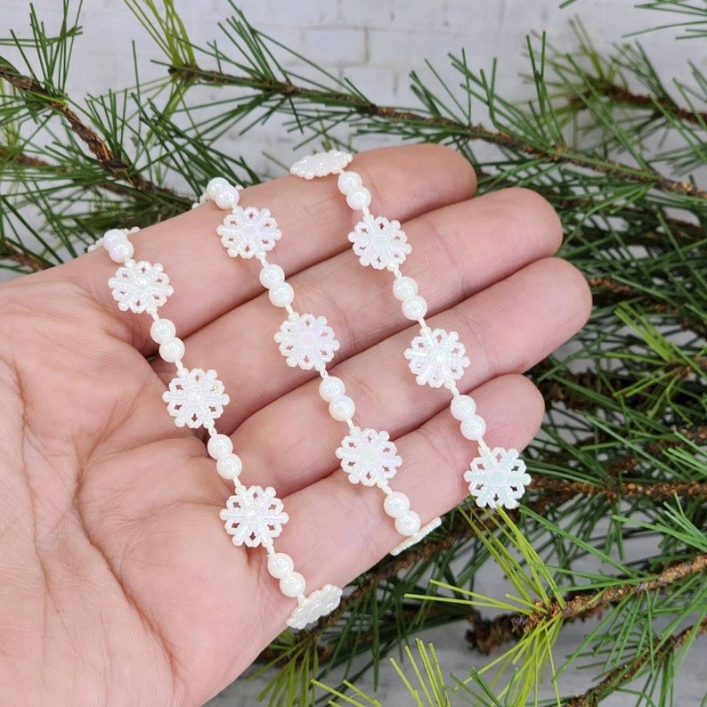 May include: White snowflake trim with pearl beads. The trim is made of white lace with a snowflake design and is strung with white pearl beads. The trim is shown on a hand against a background of green pine branches.