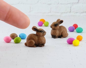 May include: Two brown rabbit figurines with painted details, surrounded by colorful Easter eggs on a white brick background.
