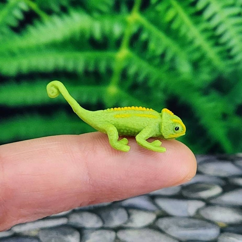May include: A small green chameleon figurine with yellow stripes, posed on a finger.