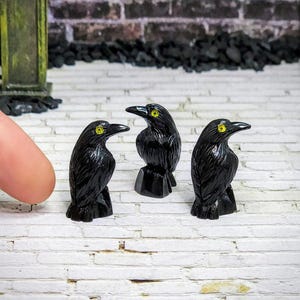 May include: Three black crow figurines with yellow eyes. The crows are carved from polished black stone and are standing on a white surface.