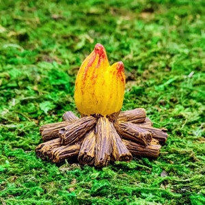 May include: A miniature campfire with a yellow and red flame made of resin. The logs are brown and the flame is sitting on top of the logs.