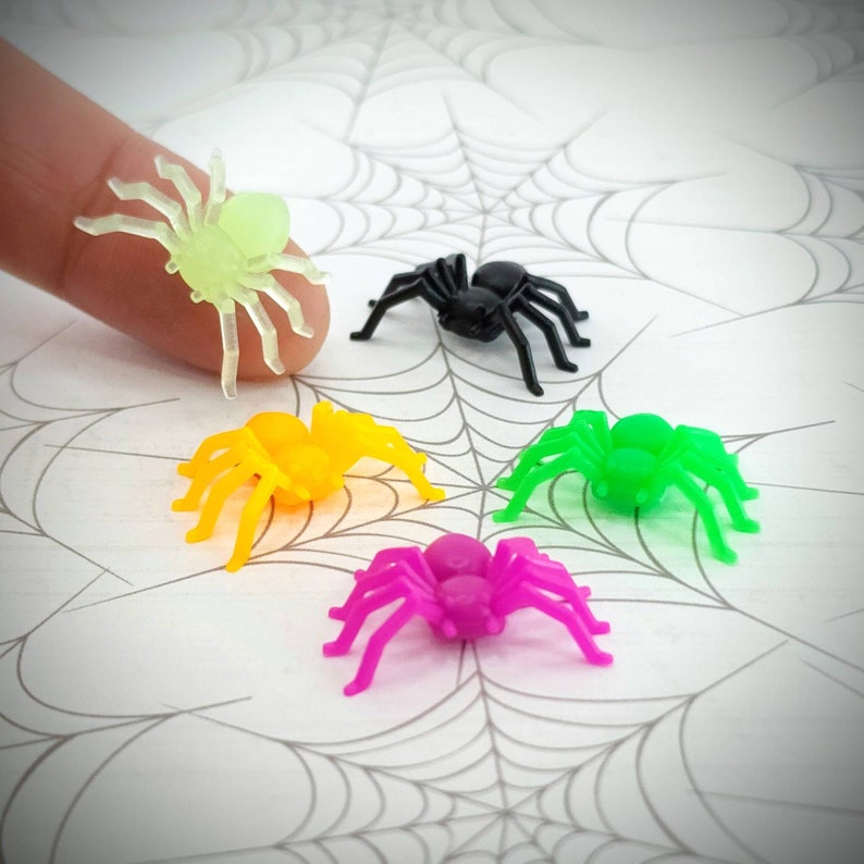 May include: Five plastic spiders in different colors: black, green, yellow, pink, and glow-in-the-dark green. The spiders are on a white background with a gray spider web pattern.