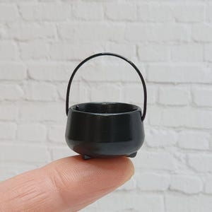 May include: A miniature black cauldron with a curved handle. The small pot has three tiny feet and a glossy finish. The cauldron is held up by a finger, against a white brick background. This item is perfect for dollhouses or Halloween decor.