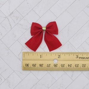 May include: A red velvet bow with a gold centre, measuring approximately 2.5 cm in width. The bow is shown next to a ruler for size reference.
