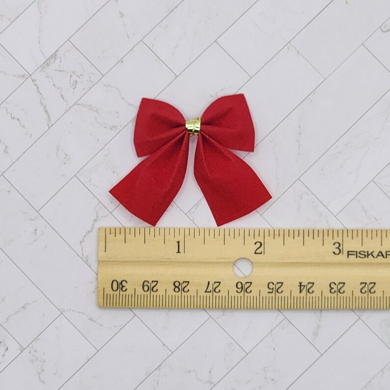 May include: A red velvet bow with a gold centre, measuring approximately 2.5 cm in width. The bow is shown next to a ruler for size reference.