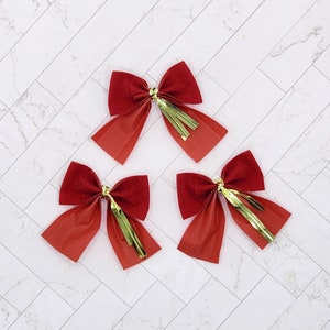 May include: Three red velvet bows with gold fringe trim. The bows are arranged in a triangular shape on a white marble background.