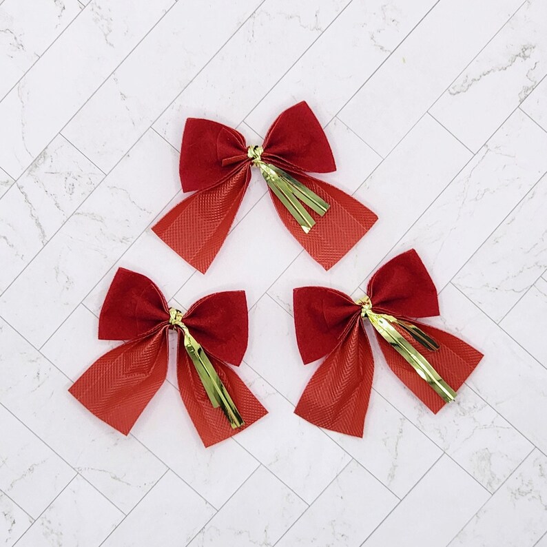 May include: Three red velvet bows with gold fringe trim. The bows are arranged in a triangular shape on a white marble background.