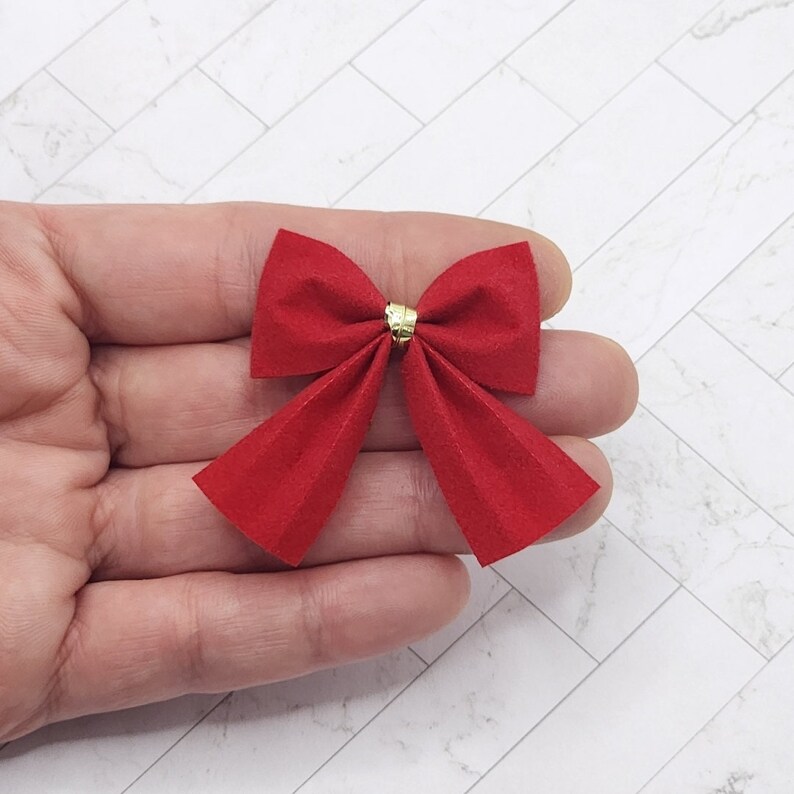 May include: A red velvet bow with a gold centre. The bow is approximately 5 cm wide and 7.5 cm long.