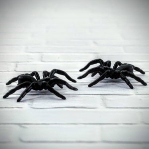 May include: Two black plastic spiders on a white brick background. The spiders have eight legs and are facing different directions.